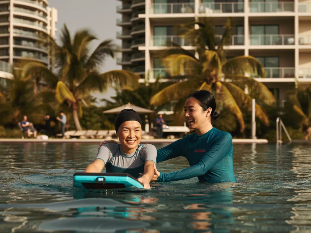 A swim coach supporting an adult learner with a kickboard in a Kuala Lumpur condominium pool.