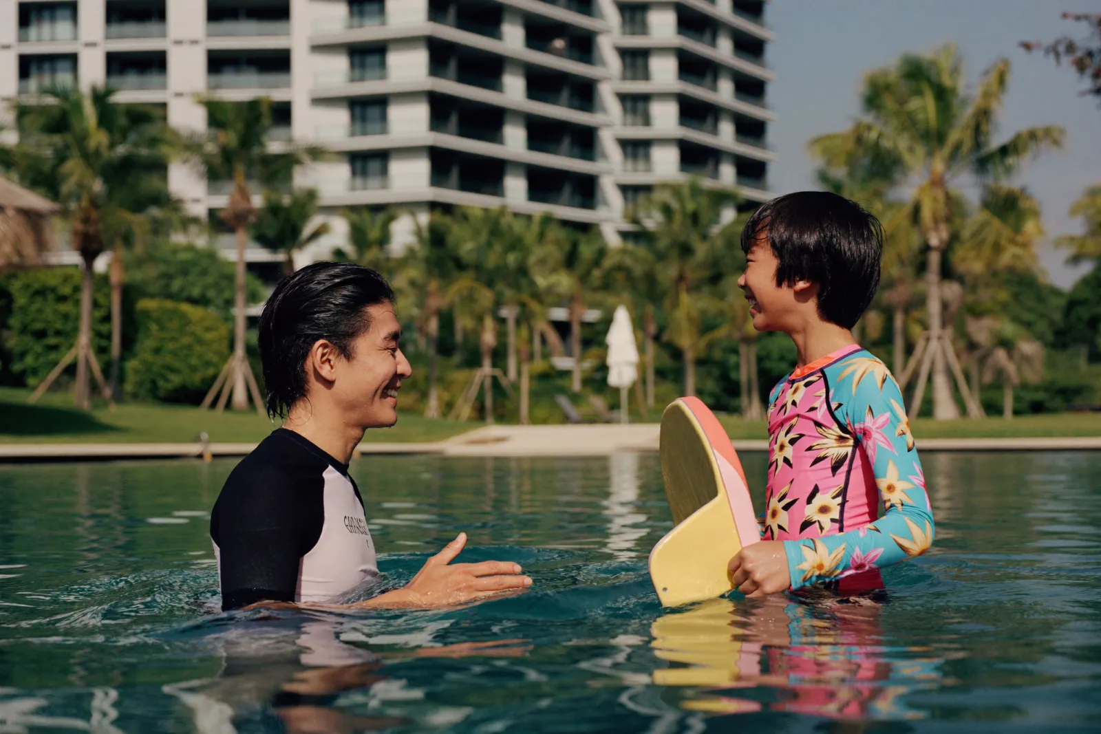 A swim coach encouraging a young child holding a kickboard in a sunlit Kuala Lumpur condominium pool.